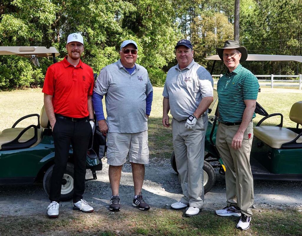 Four men standing by golf carts outdoors.